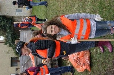 Eine Gruppe von Menschen in orangefarbenen Westen sammelt Müll auf einem Rasen. Ein Mädchen lächelt und hält einen Stock.