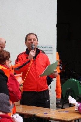Ein Mann in einem roten Jackett steht an einem Tisch, hält ein Mikrofon und ein Stück Papier in der Hand und spricht zu einer Gruppe von Menschen in Sicherheitswesten.