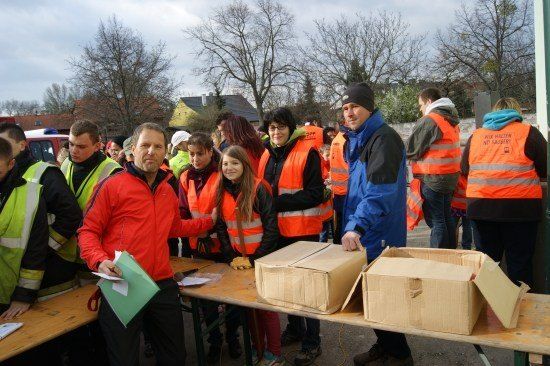 Eine Gruppe von Menschen in orangefarbenen Westen steht um einen Tisch mit Kisten, möglicherweise in Vorbereitung auf eine Veranstaltung.