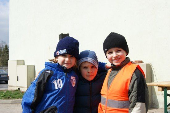 Drei Kinder stehen eng beieinander, eines trägt eine blaue Jacke mit der Nummer 10, ein anderes eine Warnweste und das dritte einen blauen Strickmütze. Sie lächeln und posieren für ein Foto.