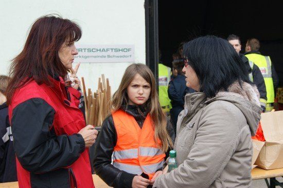 Eine Frau in einer roten Jacke spricht mit einem Mädchen in einer orangefarbenen Weste, während eine andere Frau in einer grauen Jacke zuhört. Sie stehen neben einem Tisch mit einer Tasche darauf, mit einem Schild für 'Kunstwerkstatt' im Hintergrund.