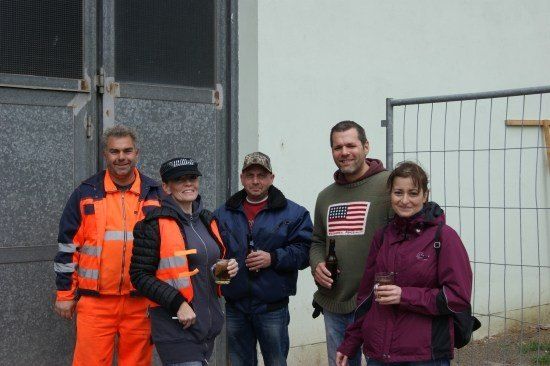 Fünf Menschen stehen vor einem Gebäude, lächeln und halten Getränke in der Hand. Der Mann auf der linken Seite trägt eine reflektierende orange Weste.