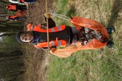 Eine Person in einer orangefarbenen Weste hält einen Stock und scheint etwas vom Boden in einem Wald zu sammeln.