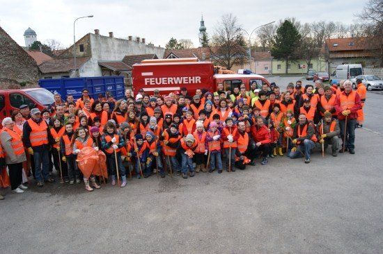Eine Gruppe von Menschen in orangefarbenen Westen posiert für ein Foto vor einem roten Feuerwehrauto. Sie halten Stöcke und einige tragen Handschuhe. Hinter ihnen stehen Gebäude, Bäume und geparkte Autos.