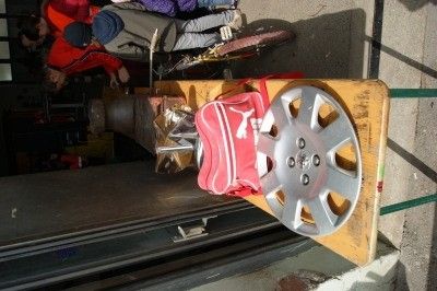 Eine silberne Autofelge liegt auf einem Holzblock mit einem roten Rucksack in der Nähe. Im Hintergrund arbeiten Menschen an einem Fahrrad.