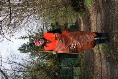 Eine Person in einem orangefarbenen Regenmantel und Hut hält einen Besen und eine Plastiktüte und steht in einem Waldgebiet.