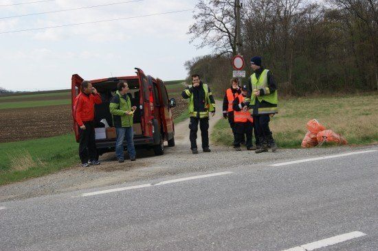Eine Gruppe von Menschen in Sicherheitswesten steht neben einem roten Van auf einer Landstraße, mit orangefarbenen Säcken auf dem Gras in der Nähe.