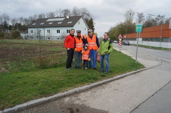 Fünf Personen, einschließlich eines Kindes, stehen auf einem Rasen neben einer Straße und tragen Warnwesten und halten Werkzeuge. Hinter ihnen ist ein Haus mit Solarpanelen auf dem Dach zu sehen.