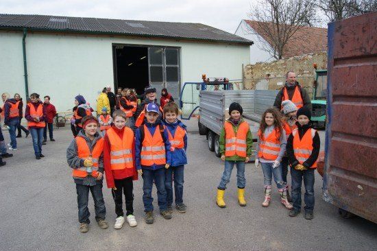 Eine Gruppe von Kindern in orangefarbenen Westen steht vor einem Gebäude mit einem Mann und einem Lastwagen im Hintergrund.