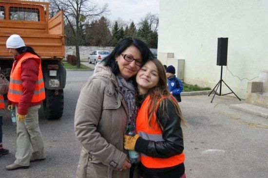 Eine Frau und ein Mädchen stehen lächelnd vor einem Gebäude mit einem Lautsprecher und einem Lastwagen im Hintergrund.