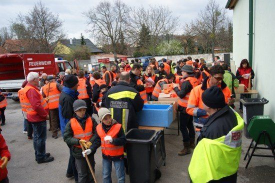 Eine Gruppe von Menschen in Sicherheitswesten versammelt sich um einen Feuerwehrwagen, einige halten Besen und andere stehen in der Nähe eines Tisches mit Lagerboxen. Eine Frau liest ein Papier.