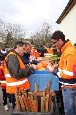 Eine Gruppe von Menschen in orangenen Westen steht um einen Tisch mit Stöcken und Essen, möglicherweise in Vorbereitung auf eine Veranstaltung.