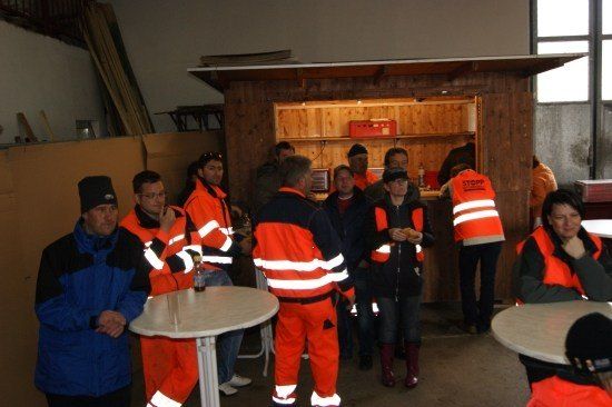 Eine Gruppe von Menschen in Warnwesten steht um einen Tisch in einem Raum. Einige stehen, andere sitzen. Hinter ihnen befindet sich ein Holztresen mit Regalen.