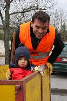 Ein Mann mit oranger Warnweste und gelben Handschuhen lächelt, während er ein Kind in einem gelben Wagen auf der Straße hält.