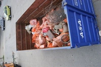 Ein Blick von oben auf einen blauen Müllcontainer, gefüllt mit verschiedenen Abfällen, einschließlich Plastiktüten und anderen Gegenständen. Ein grünes Auto ist in der Nähe geparkt.