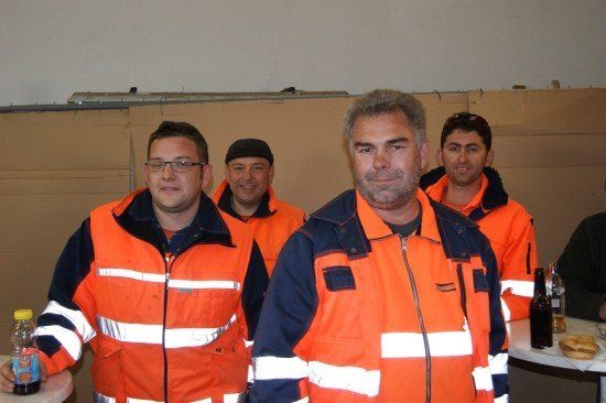 Fünf Männer in orangefarbenen Sicherheitswesten stehen zusammen und lächeln für ein Foto. Einer hält eine Flasche, und andere haben Getränke auf einem Tisch.