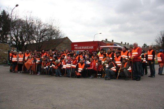 Eine Gruppe von Menschen in orangefarbenen Westen posiert für ein Foto vor einem Feuerwehrauto. Die Szene ist im Freien mit Gebäuden und Bäumen im Hintergrund.