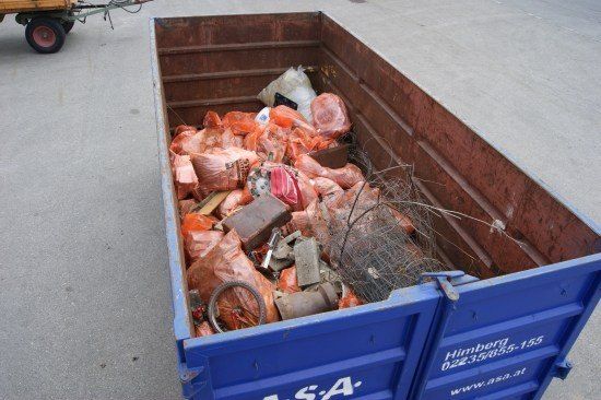 Ein blauer Müllcontainer gefüllt mit verschiedenen Abfällen, einschließlich Plastiktüten, einem Fahrradrad und anderen Gegenständen, mit einem Firmenlogo an der Seite.