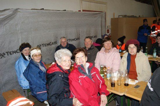 Eine Gruppe älterer Menschen sitzt an einem Tisch in einem Raum, einige tragen Brillen und Hüte. Flaschen und Gläser stehen auf dem Tisch. Ein weißer Banner mit schwarzem Text im Hintergrund.
