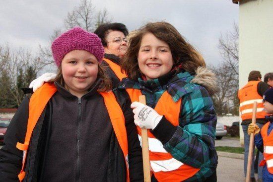 Zwei junge Mädchen in orangefarbenen Westen und Handschuhen lächeln und posieren für ein Foto mit einem Holzstock. Dahinter lächelt auch eine Frau mit Brille. Sie befinden sich in einem Außenbereich mit Bäumen und einem Gebäude.