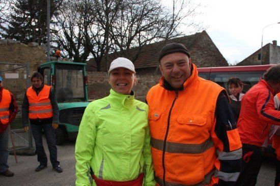 Ein Mann und eine Frau in Warnwesten stehen lächelnd nebeneinander vor einem Traktor und einem Bus. Dahinter stehen mehrere Personen, von denen eine einen blauen Hut trägt.