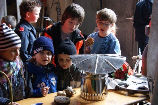Eine Gruppe von Kindern versammelt sich um einen Tisch und untersucht ein Metallobjekt. Einige tragen Winterkleidung, und ein Kind isst.