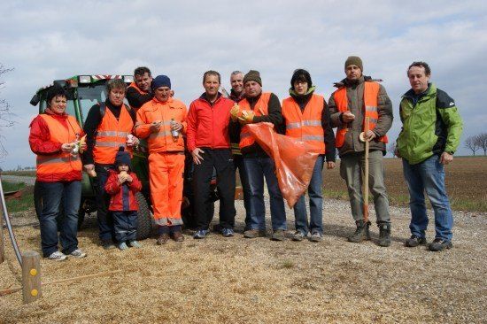 Eine Gruppe von Menschen in orangefarbenen Westen und Winterkleidung steht auf einem Feld, einige halten Essen und andere halten orange Taschen. Ein Traktor ist im Hintergrund sichtbar.