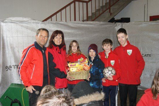 Eine Gruppe von Menschen in roten Jacken, darunter zwei Erwachsene und vier Kinder, stehen zusammen. Ein Kind hält einen Korb mit Snacks und ein anderes ein Fußball. Sie lächeln und scheinen für ein Foto zu posieren.