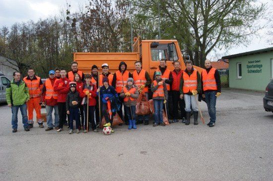 Eine Gruppe von Menschen in orangefarbenen Westen und Handschuhen, einige halten Besen und Taschen, posiert für ein Foto vor einem großen orangefarbenen Lastwagen.