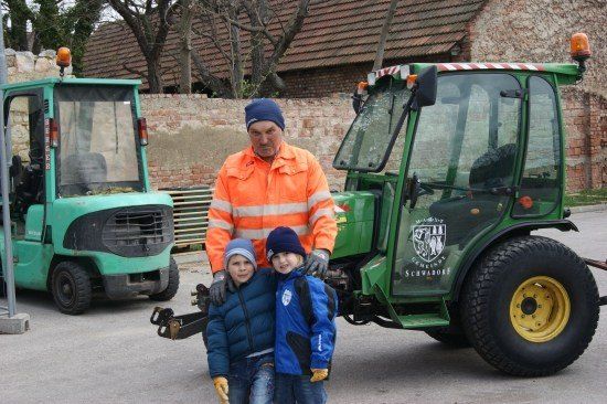 Ein älterer Mann und zwei Kinder stehen vor einem grünen Traktor, mit einer Ziegelmauer und einem weiteren Traktor im Hintergrund.