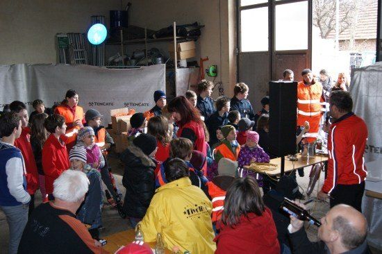 Eine Gruppe von Menschen in Sicherheitswesten versammelt sich in einem Raum, einige tragen Mützen und andere halten Flaschen. Ein Mann in einer roten Jacke spricht zu der Menge.