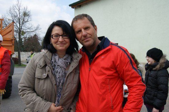 Ein Mann und eine Frau stehen eng beieinander und lächeln für ein Foto vor einem Gebäude. Dahinter ist eine Person in einer Warnweste zu sehen, und eine weitere Person ist teilweise verdeckt.
