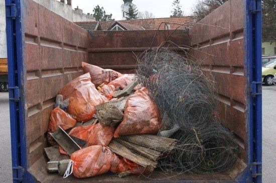Ein blauer Container gefüllt mit verschiedenen Abfallmaterialien, darunter orange Plastiksäcke, Holzstücke und gewickelte Drähte.
