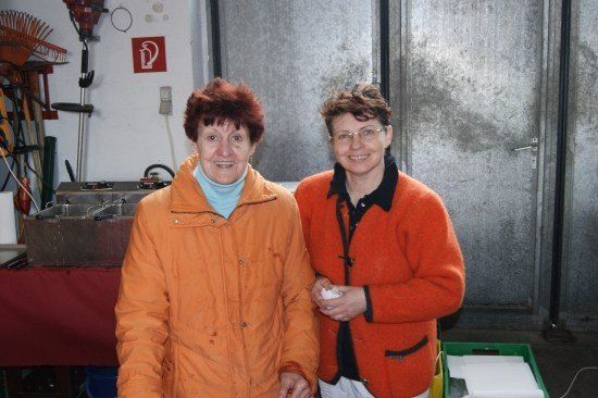 Zwei Frauen in orangefarbenen Jacken stehen nebeneinander in einem Raum mit einem roten Tisch und einem Feuerlöscher-Schild an der Wand.