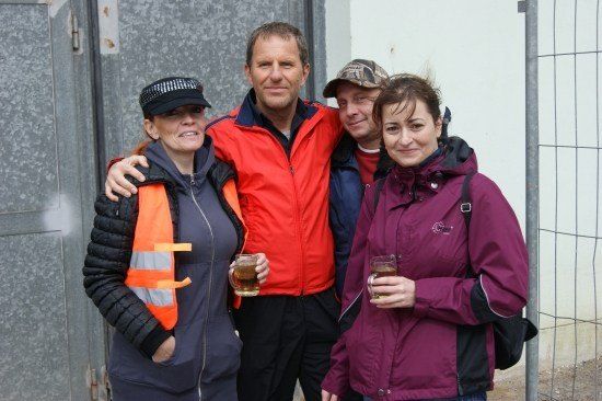 Vier Personen, zwei Frauen und zwei Männer, stehen zusammen draußen. Sie alle lächeln und halten Gläser mit Bier. Der Mann in der Mitte trägt eine rote Jacke. Hinter ihnen ist eine graue Wand mit einem Metallzaun.