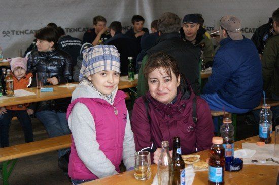 Eine Gruppe von Menschen sitzt an einem Tisch in einem Zelt, lächelt und posiert für ein Foto. Es gibt Flaschen, Gläser und Essen auf dem Tisch. Das Zelt hat eine weiße Wand mit etwas Text darauf.