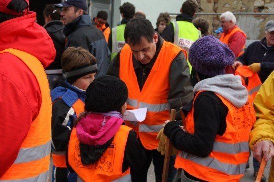 Eine Gruppe von Menschen in Warnwesten ist um einen Mann versammelt, der Anweisungen zu geben scheint. Die Menschen tragen Winterkleidung und einige halten Stöcke.