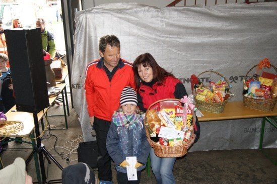 Ein Mann und eine Frau in roten Jacken stehen mit einem Kind, das einen Korb mit Essen hält. Dahinter steht ein Tisch mit zwei Körben voller Snacks. Im Hintergrund bedeckt eine weiße Plane eine Tür.