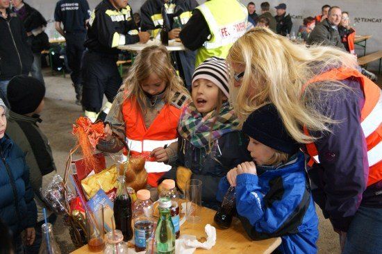 Eine Gruppe von Kindern und Erwachsenen versammelt sich um einen Tisch mit verschiedenen Snacks und Getränken. Die Kinder tragen orange Westen. Eine Frau in einer blauen Jacke schaut die Kinder an.