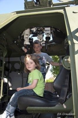 Zwei Kinder sitzen in einem Militärfahrzeug, eines hält eine Kamera und das andere schaut darauf. Das Fahrzeug hat eine grüne Farbe und einen schwarzen Sitz.