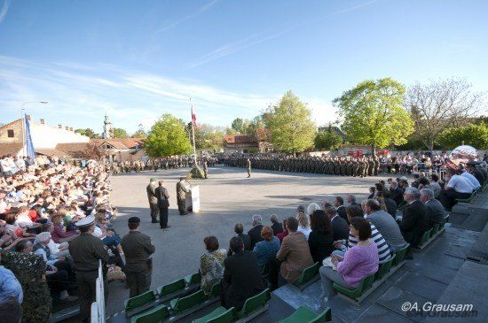 Eine große Menschenmenge versammelt sich für eine formelle Veranstaltung im Freien. In der Mitte steht ein Podium, dahinter ein Fahnenmast. Die Teilnehmer sitzen auf grünen Bänken, einige stehen. In der Ferne steht eine Reihe von Soldaten in Aufmerksamkeit.
