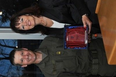 Ein Mann und eine Frau lächeln und posieren für ein Foto, während sie eine Trophäe in einer blauen Schachtel halten. Der Mann trägt eine Militäruniform und die Frau trägt einen schwarzen Blazer.