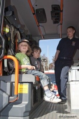 Drei Kinder sitzen in einem Krankenwagen, eines trägt ein grünes Shirt und einen Hut, während ein Feuerwehrmann in der Nähe steht und möglicherweise etwas erklärt.