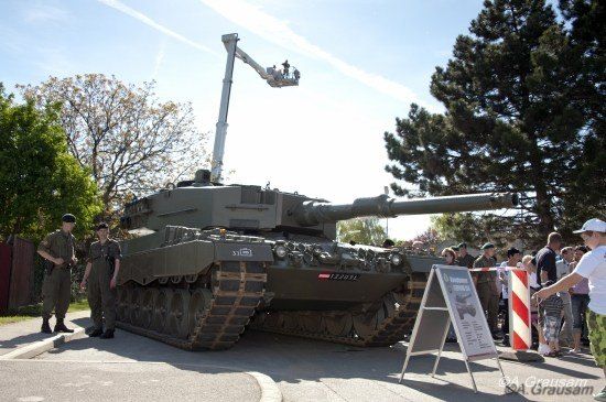 Ein großer Militärpanzer wird im Freien ausgestellt, wobei zwei Soldaten in der Nähe stehen und eine Menschenmenge zusieht.