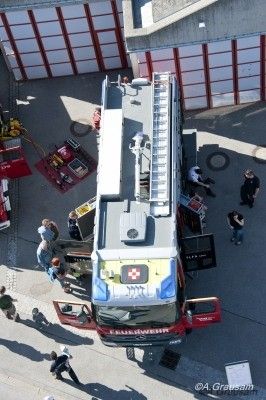 Eine Draufsicht auf einen Feuerwehrwagen mit offenen Türen, umgeben von Menschen auf einem Parkplatz. Der Feuerwehrwagen hat ein rotes Kreuz an der Seite und eine Leiter auf dem Dach.