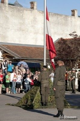 Zwei Soldaten in Uniform hissen eine Flagge vor einer Menschenmenge, mit einem Gebäude und Bäumen im Hintergrund.