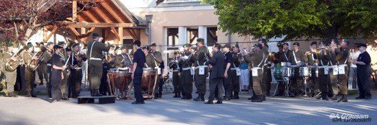Ein Militärorchester in Uniform spielt auf der Straße mit Blech- und Schlaginstrumenten unter einem Baum.