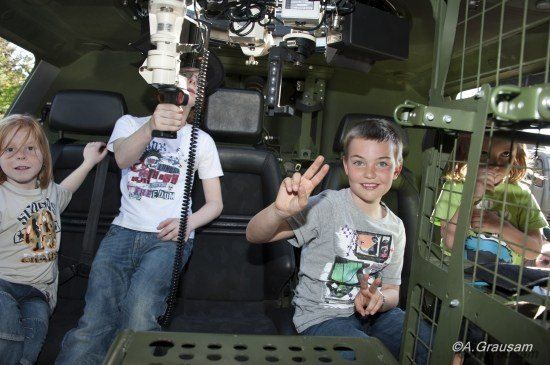 Vier Kinder sitzen in einem Militärfahrzeug, zwei Jungen machen Friedenszeichen, einer hält eine Kamera und ein Mädchen schaut aus dem Fenster.