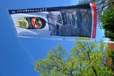 Ein großes Banner mit einem Bild eines Militärfahrzeugs und Text in einer fremden Sprache weht im Wind gegen einen blauen Himmel mit grünen Bäumen.