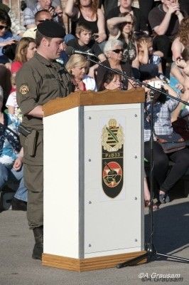 Ein Mann in Uniform steht an einem Rednerpult mit Mikrofon und spricht vor einem Publikum. Das Pult hat ein Logo und der Mann trägt einen Hut und Stiefel.